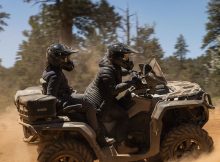 Two riders on an ATV driving through a dusty forest terrain.